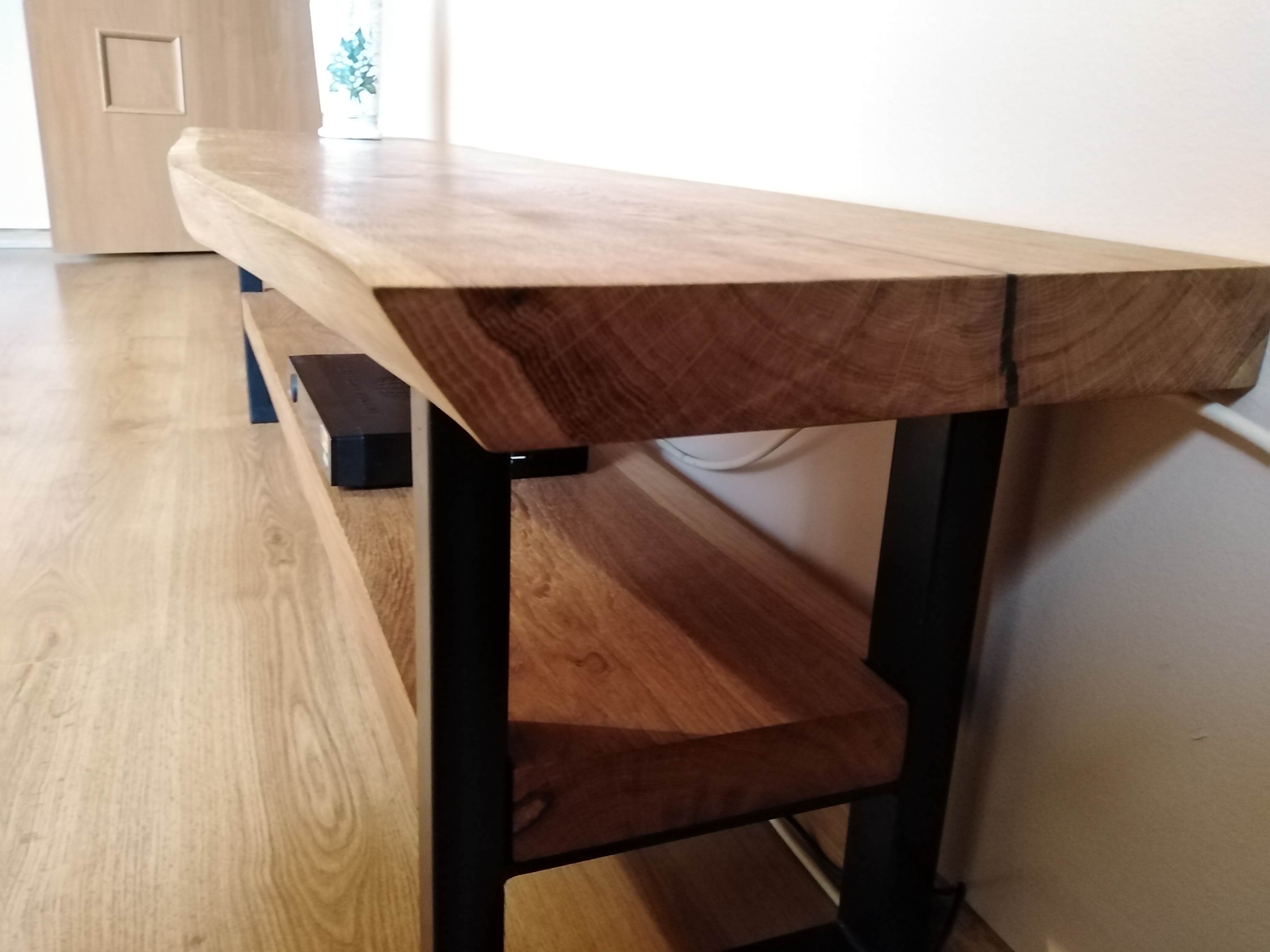 Drewniana konsola RTV z blatem o nieregularnej krawędzi i czarnymi metalowymi nogami, widoczna na tle drewnianej podłogi i jasnej ściany.