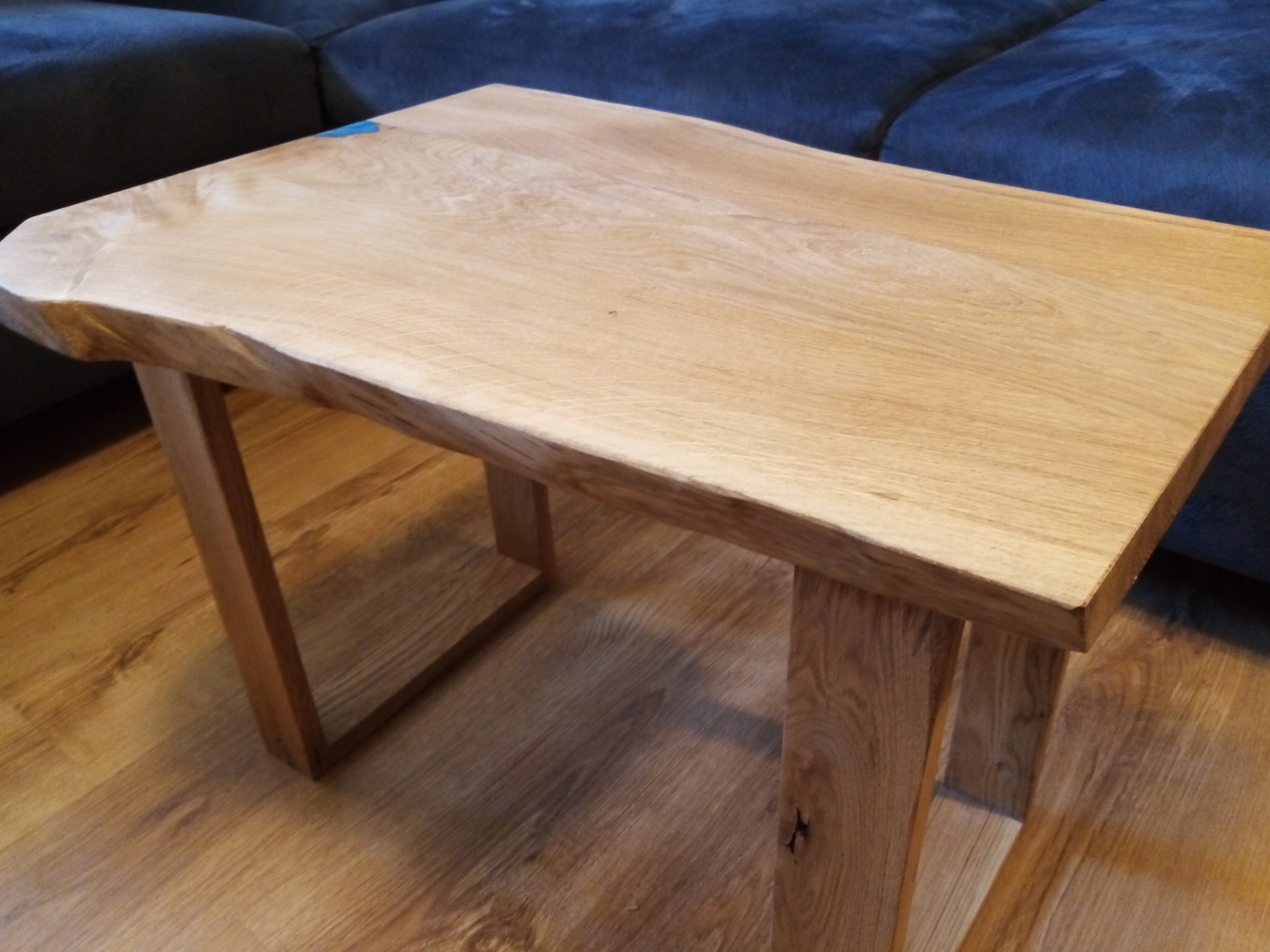 Niski stolik kawowy z blatem z litego drewna o naturalnych krawędziach, ustawiony na drewnianej podłodze, z fragmentem granatowej sofy w tle.