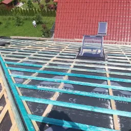 Konstrukcja dachu z drewnianych łat pokrytych czarną membraną, widoczne okna dachowe, w tle czerwona dachówka i zielony trawnik z ludźmi.