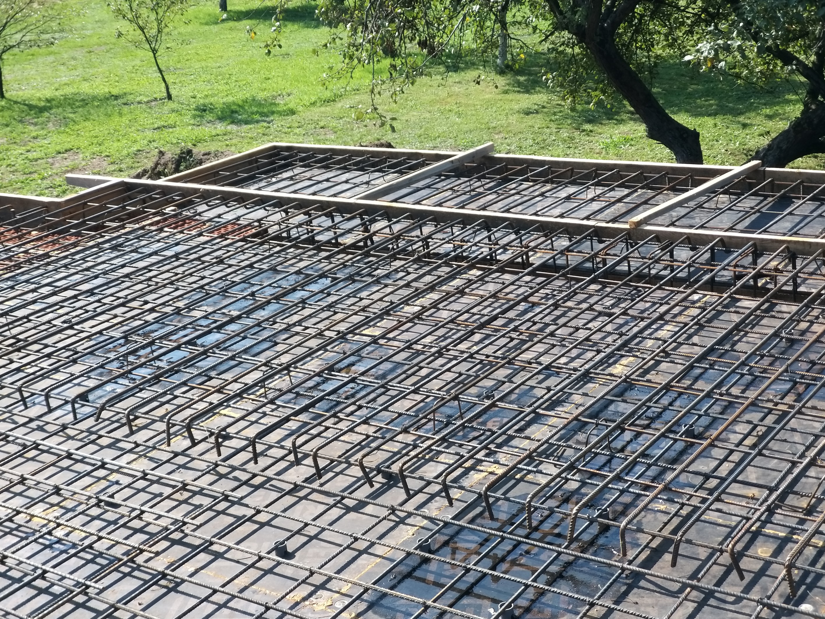 Siatka zbrojeniowa ułożona na czarnej folii, przygotowana do zalania betonem pod fundamenty, widoczne drewniane szalunki i zielona trawa w tle.