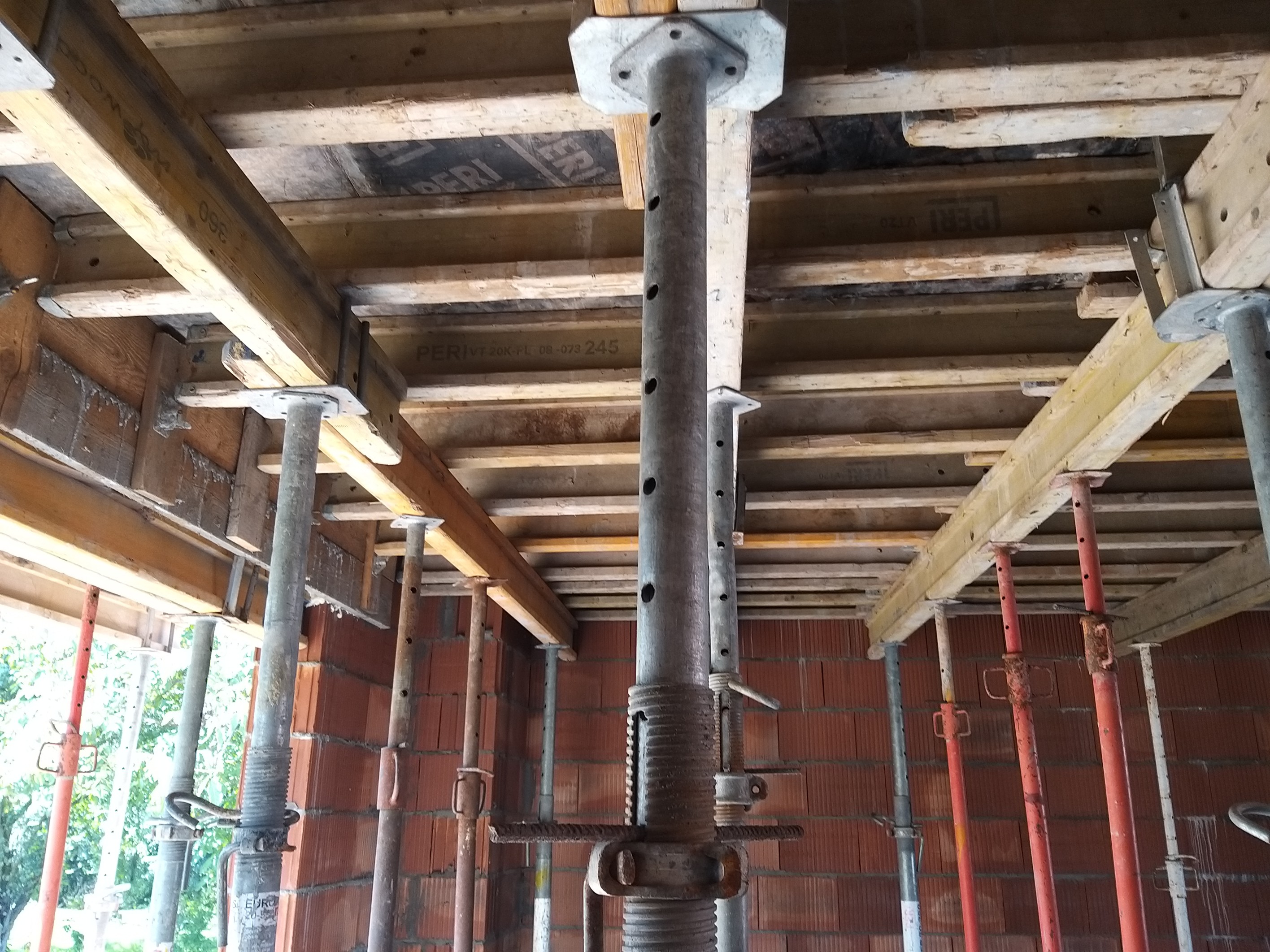 Szare stemple budowlane podtrzymujące drewniany szalunek stropu z widocznymi deskami i płytami szalunkowymi w trakcie budowy domu z czerwonej cegły.