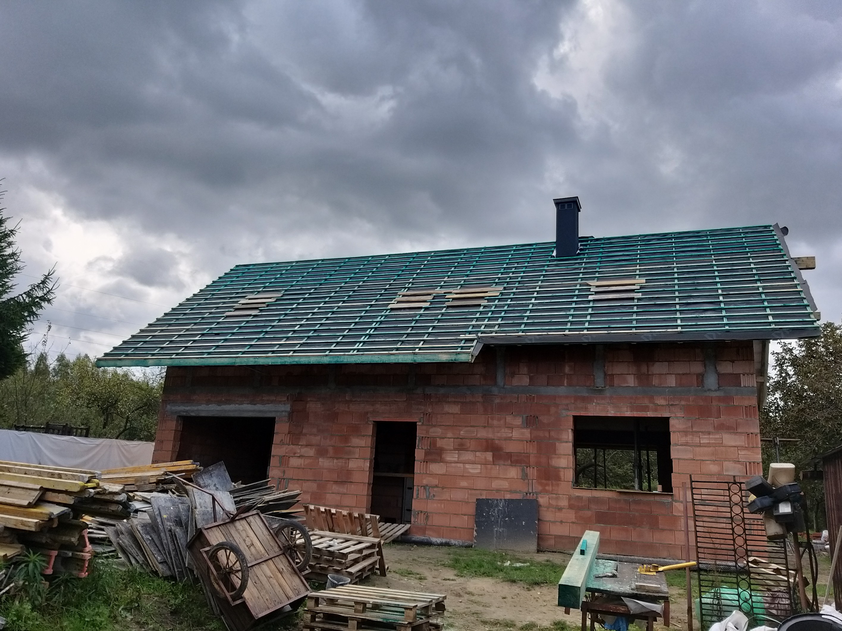 Surowy stan domu z czerwonej cegły pod ciemnym, zachmurzonym niebem, z przygotowaną konstrukcją dachu i czarnym kominem, widoczne materiały budowlane i palety na pierwszym planie.