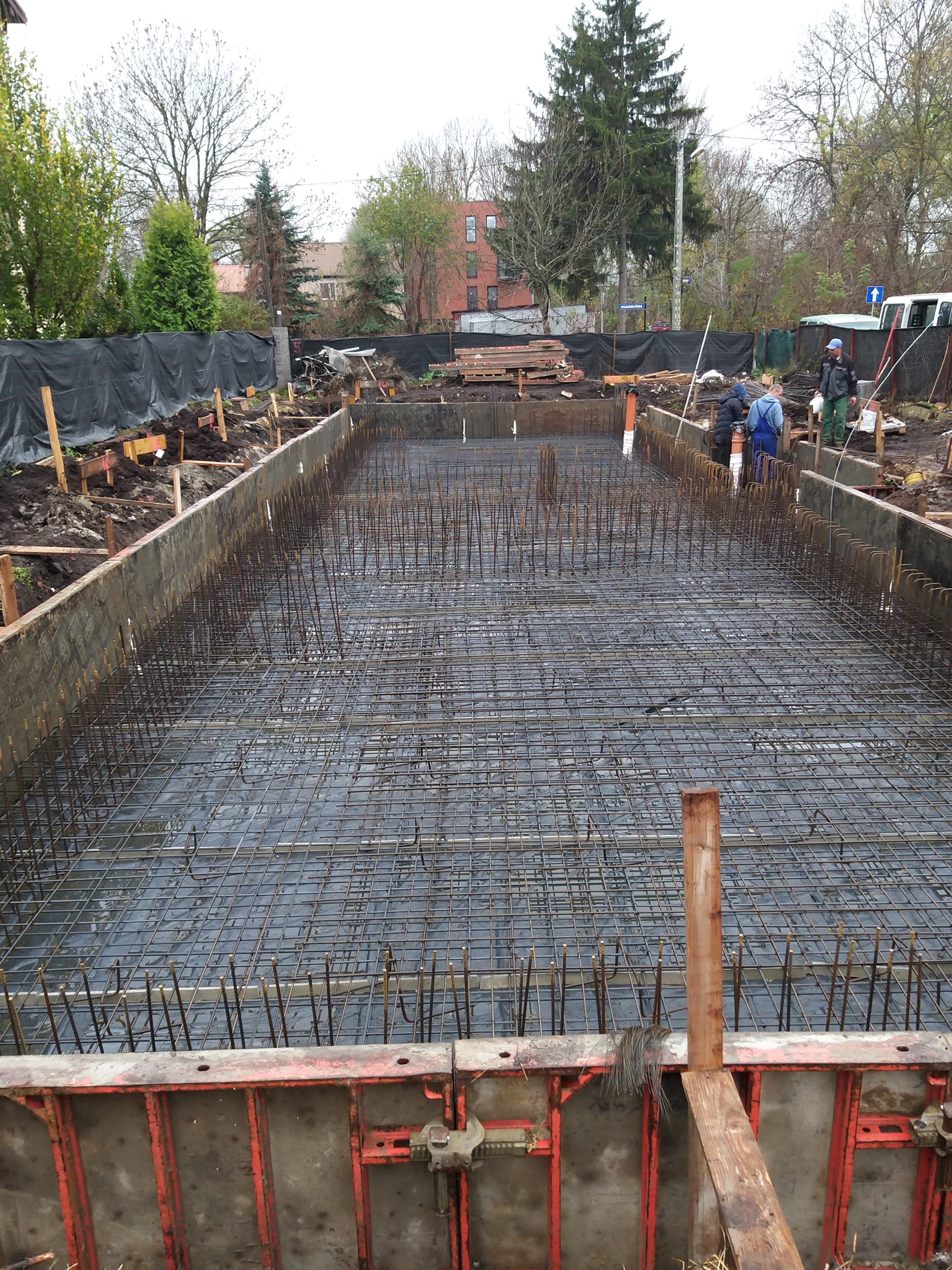 Zbrojenie fundamentu pod budynek w Krakowie, widoczne szalunki i siatka zbrojeniowa, w tle pracownicy budowlani i otoczenie placu budowy.