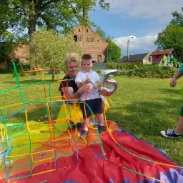 Animator z dzieckiem na trawie, z kolorowym tunelem z rurek i balonem w kształcie cyfry 5, w tle budynek z cegły i drzewa.