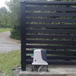 Zewnętrzny napęd automatyczny do bramy przesuwnej Nice Robus zamontowany na betonowej podstawie przy czarnej metalowej bramie z poziomymi panelami, widok z boku na tle zieleni.