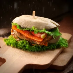 Apetyczny, warstwowy burger z grillowanym kurczakiem, pomidorem, marchewką i sałatą, spięty wykałaczką, prezentowany na drewnianej desce do krojenia w scenerii padającego cukru pudru.