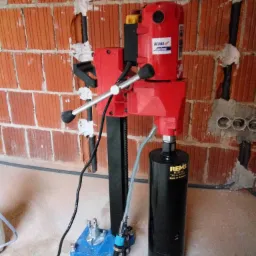 Czerwona wiertnica diamentowa Dedra Maxima z czarnym wiertłem REMS na nieotynkowanej ścianie z czerwonej cegły z widocznymi kablami i rurami.