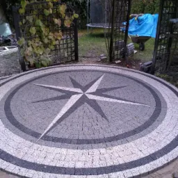 Okrągły plac z kostki granitowej ułożonej we wzór kompasu, obrzeże w trakcie wykańczania, w tle altana, trampolina i wózek budowlany.
