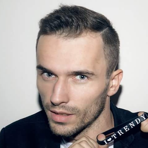 Portret młodego mężczyzny z krótką fryzurą i lekkim zarostem, trzymającego w dłoni czarny przedmiot z napisem 'I-TRENDY', na jasnym tle.