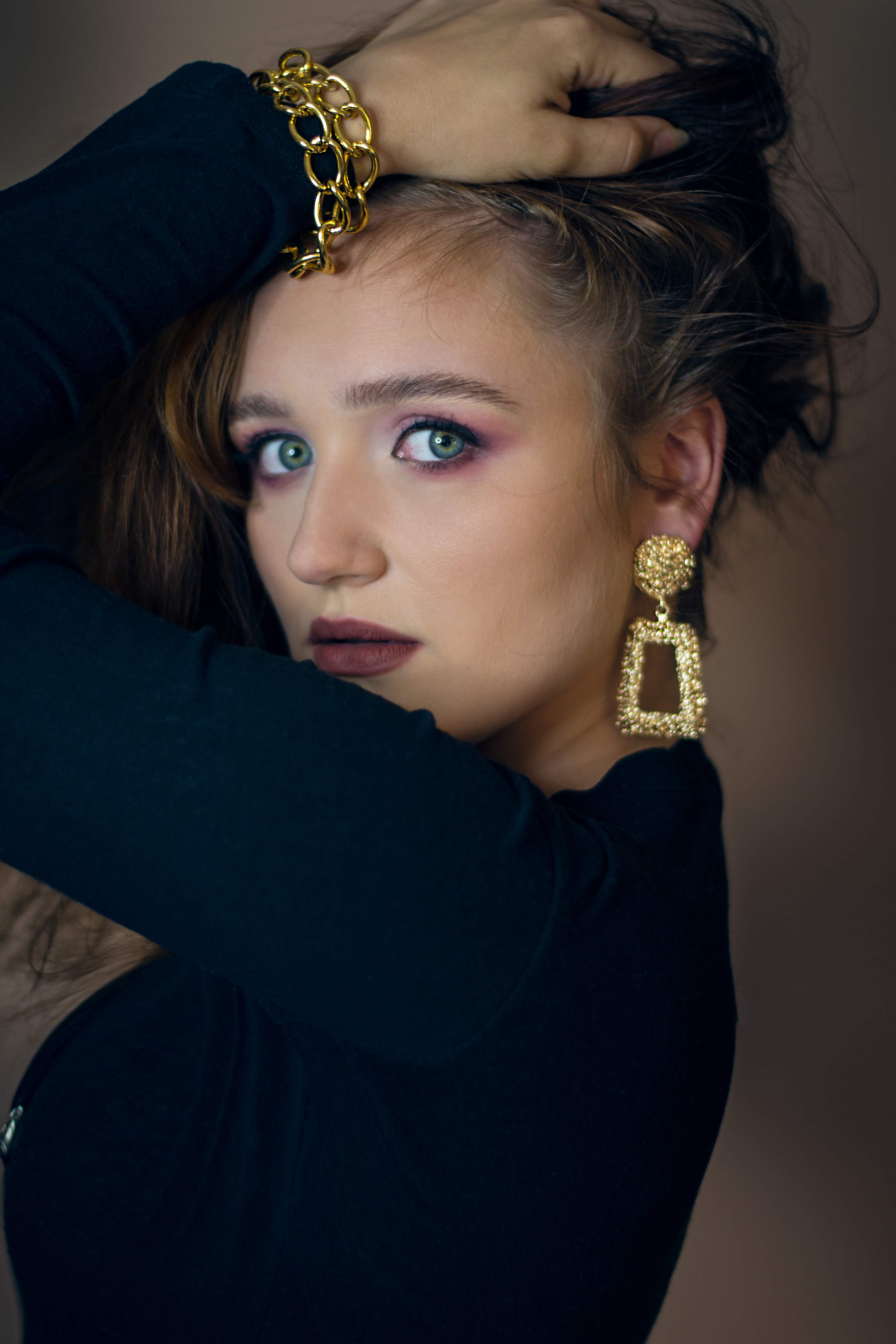 Portret kobiety z makijażem w odcieniach fioletu, złota biżuteria na szyi i uszach, dłoń we włosach, ciemne ubranie na beżowym tle.