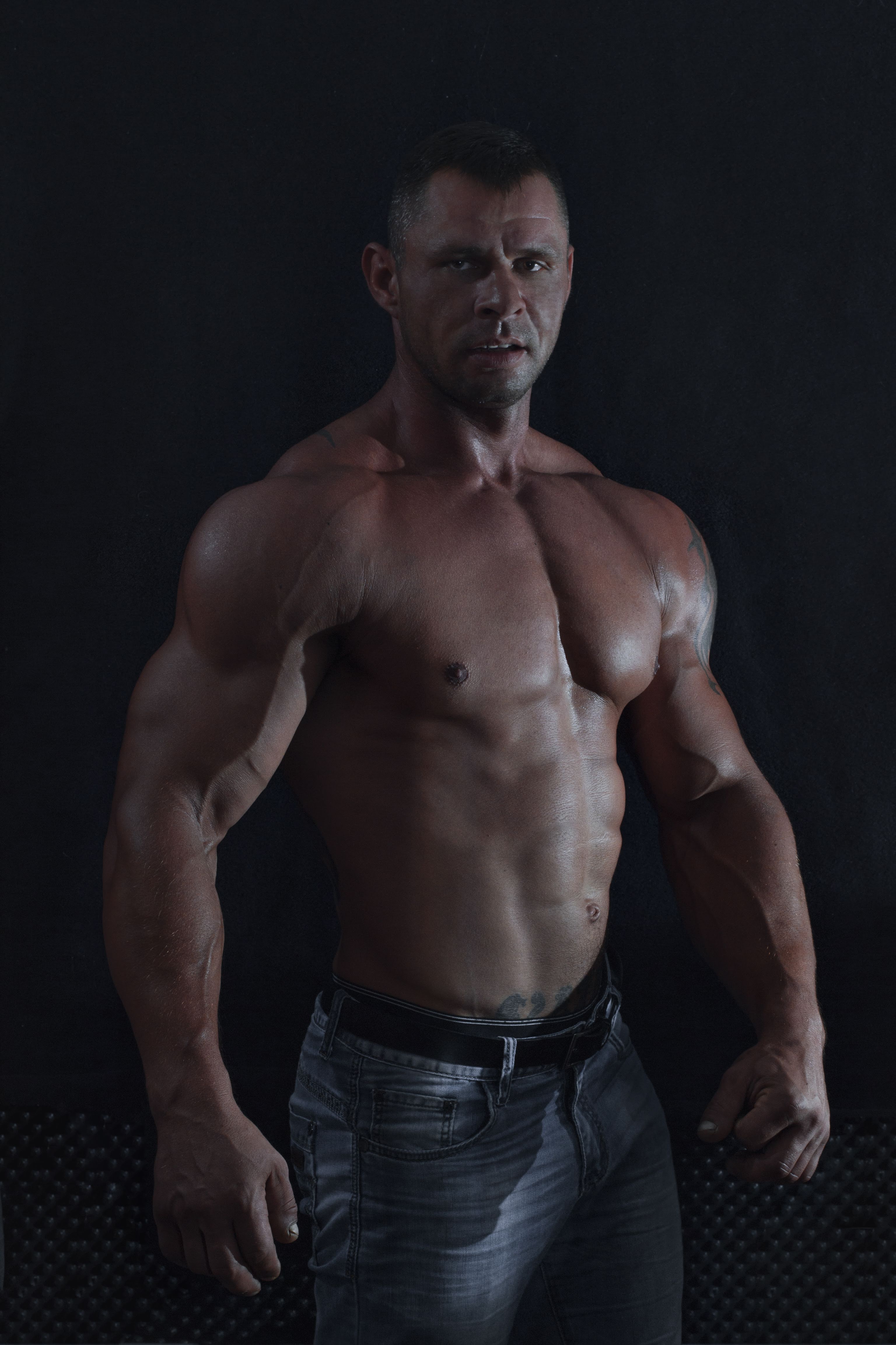 Mężczyzna o muskularnej sylwetce, z widocznymi żyłami na ramionach, pozuje na ciemnym tle, ubrany w dżinsy i z tatuażami na ciele.