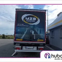 Tylna ściana naczepy ciężarówki z nadrukowanym logo firmy transportowej MZB, numerem telefonu i adresem e-mail, widoczna na parkingu w słoneczny dzień.