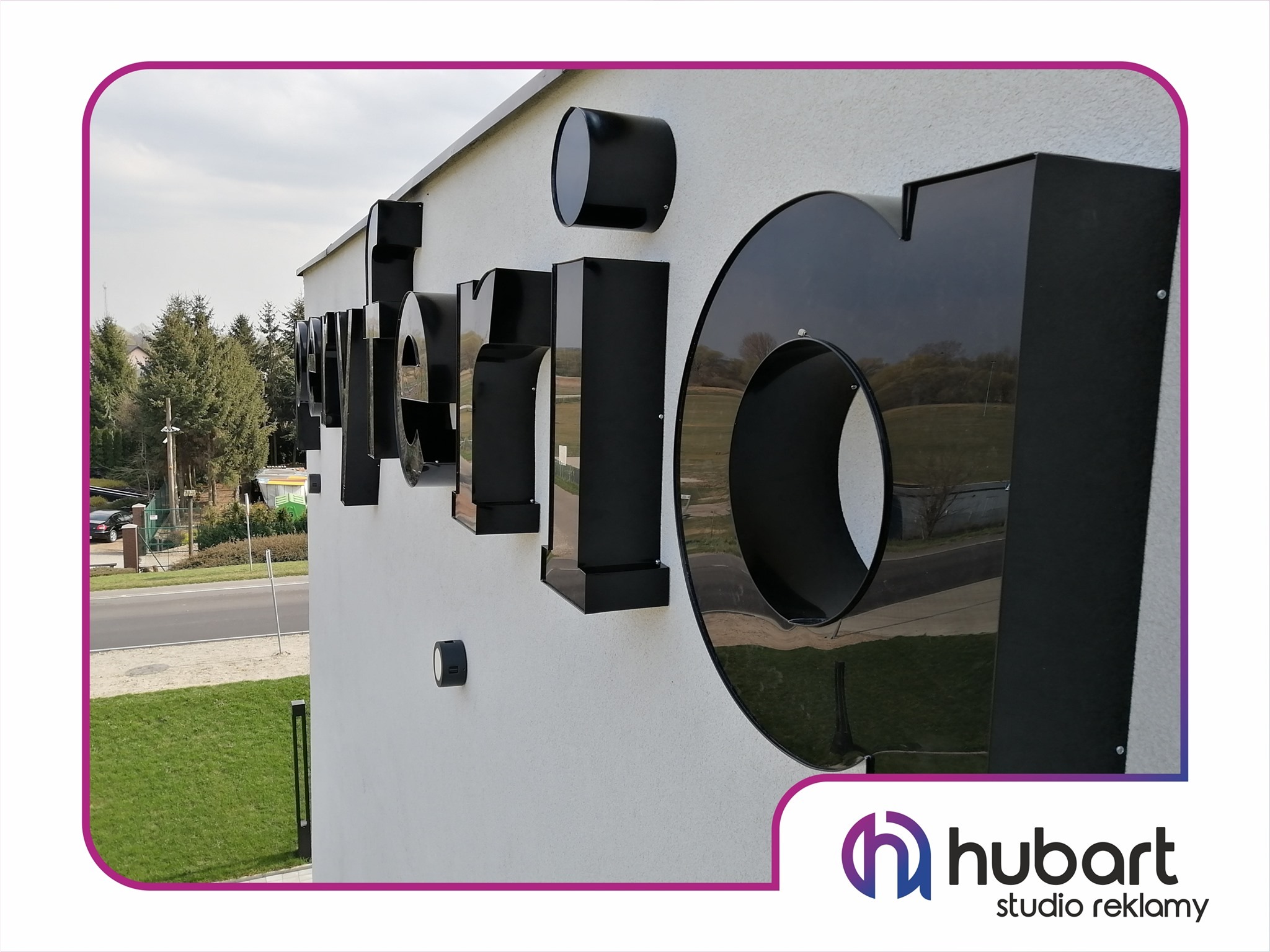 Elewacja budynku z dużym, czarnym, przestrzennym napisem reklamowym 'hubart studio reklamy', widoczne drzewa i ulica w odbiciu liter.