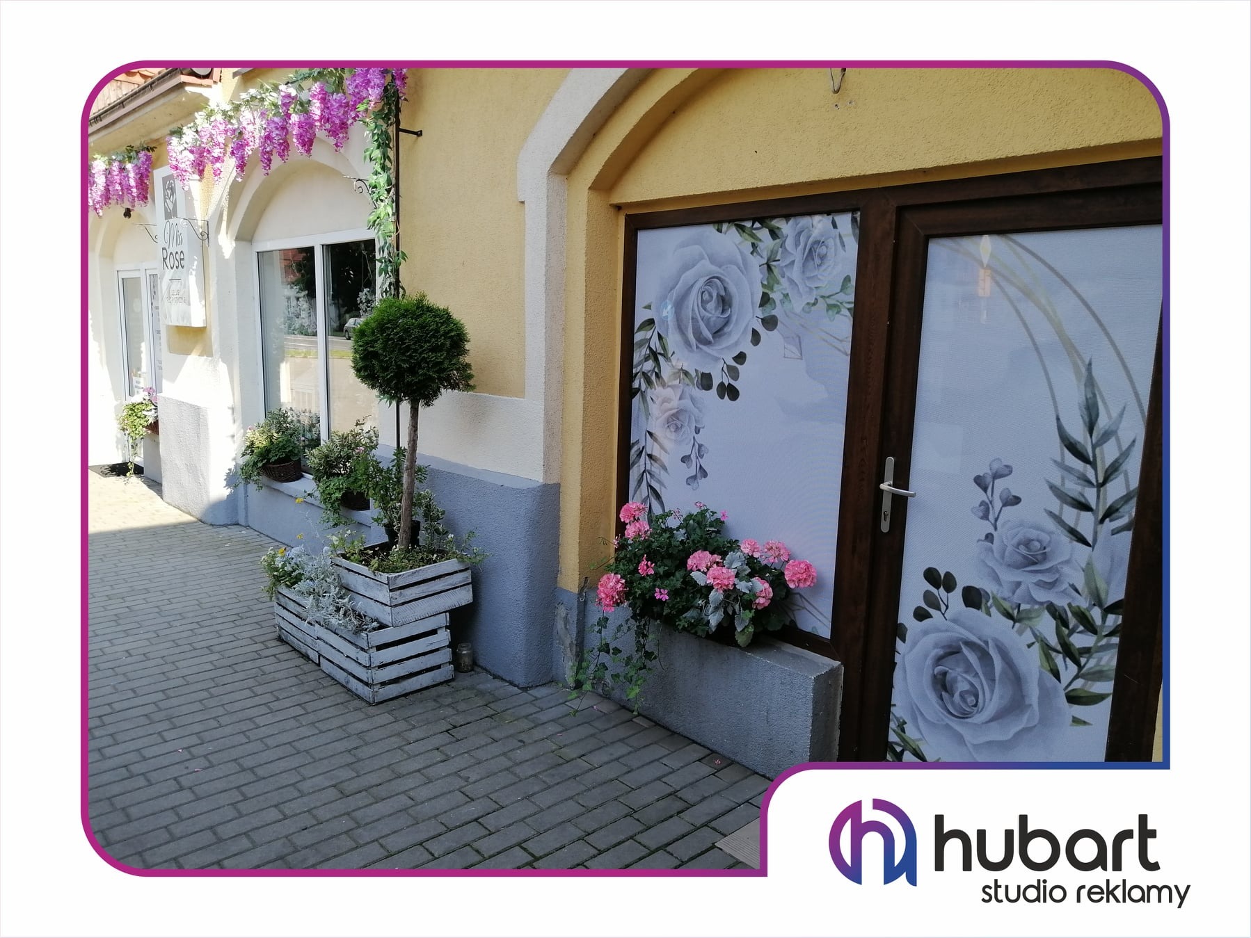 Okna lokalu ozdobione folią z motywem kwiatowym, z zewnątrz donice z kwiatami i krzewami.