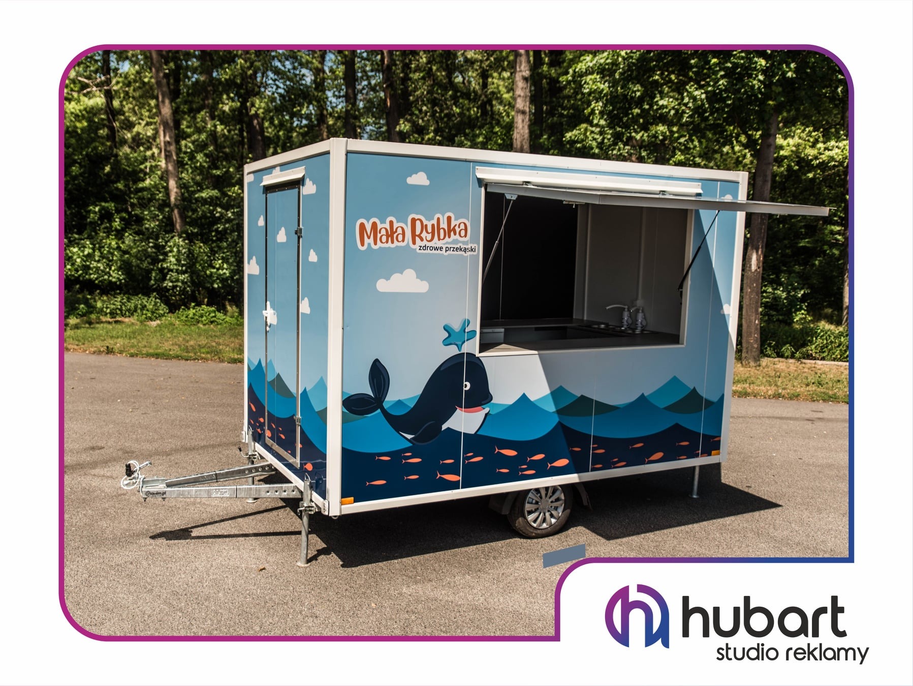 Przyczepa gastronomiczna z nadrukiem grafiki morskiej, w tym wieloryba i ryb, z otwartym okienkiem sprzedażowym, stojąca na parkingu z drzewami w tle.