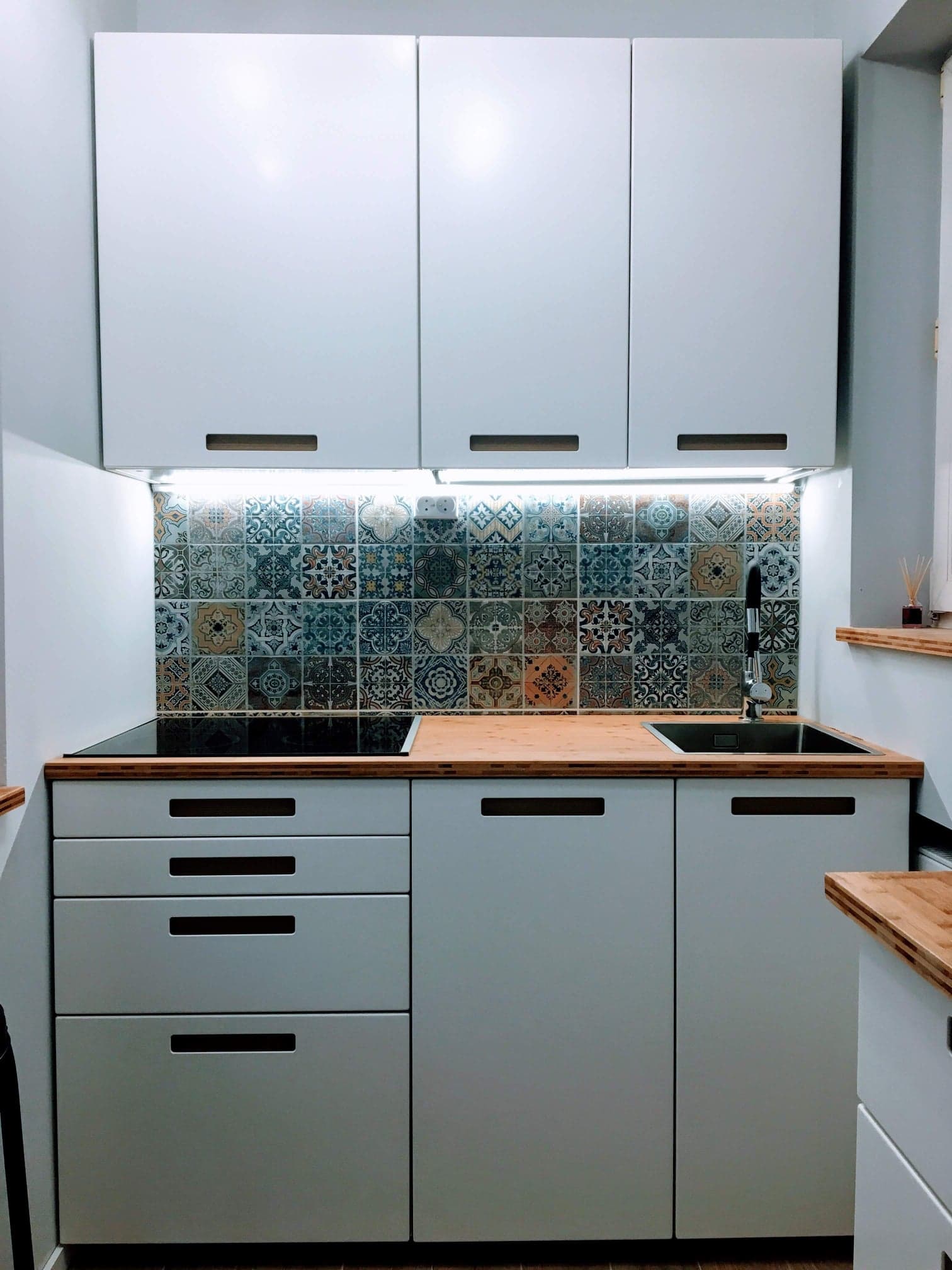 Kompaktowa, jasnoszara kuchnia z drewnianym blatem i dekoracyjnymi, wielokolorowymi płytkami nad blatem, oświetlona listwą LED