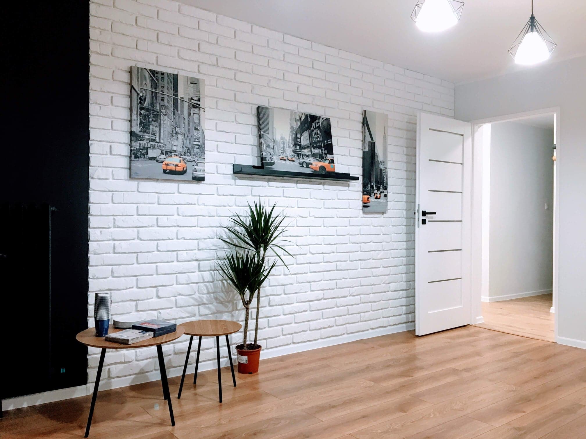 Wnętrze pokoju z białą ścianą z cegieł, trzema obrazami z motywem Nowego Jorku, drewnianą podłogą, białymi drzwiami i dwiema lampami sufitowymi.