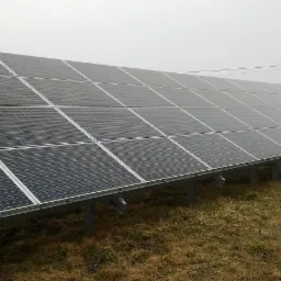 Rząd paneli fotowoltaicznych zamontowanych na metalowej konstrukcji na trawiastym terenie, widoczne słupy energetyczne w oddali, pochmurne niebo.