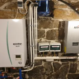 Instalacja dwóch urządzeń: biały moduł Ecodan z wyświetlaczem i zielonym akcentem oraz srebrny moduł Afore, oba zamontowane na ścianie z kamienia, z widocznymi rurami i przewodami.