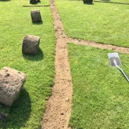 Przygotowany teren pod trawnik z rolki: wyznaczone ścieżki, wykopana ziemia, zwinięte rolki trawy oraz szpadel w tle.