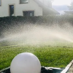 Automatyczne zraszacze nawadniające trawnik na tle domu jednorodzinnego, widoczna biała lampa ogrodowa.