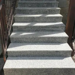 Zewnętrzne schody z jasnoszarego granitu z kutą balustradą prowadzące do drewnianych drzwi wejściowych.