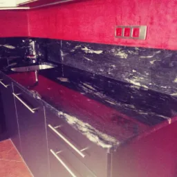 Czarny blat kuchenny z marmurkowym wzorem, zamontowany na ciemnych szafkach z metalowymi uchwytami, widoczny zlew i bateria, intensywnie czerwona ściana w tle.