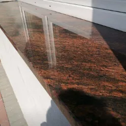 Polerowany parapet zewnętrzny z brązowo-czarnego granitu, odbijający okno, zamontowany na białej ścianie z widocznym fragmentem chodnika.