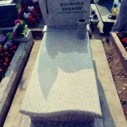 Jasnoszary, granitowy pomnik z wyrytym krzyżem i inskrypcją, otoczony kwiatami i zniczami na cmentarzu.