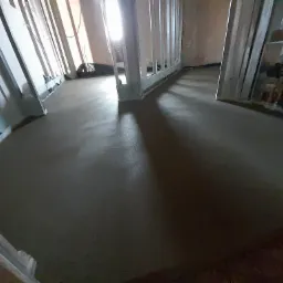 Świeżo wylana, gładka posadzka w trakcie schnięcia, widoczne profile oddzielające strefy, instalacja ogrzewania podłogowego po prawej.