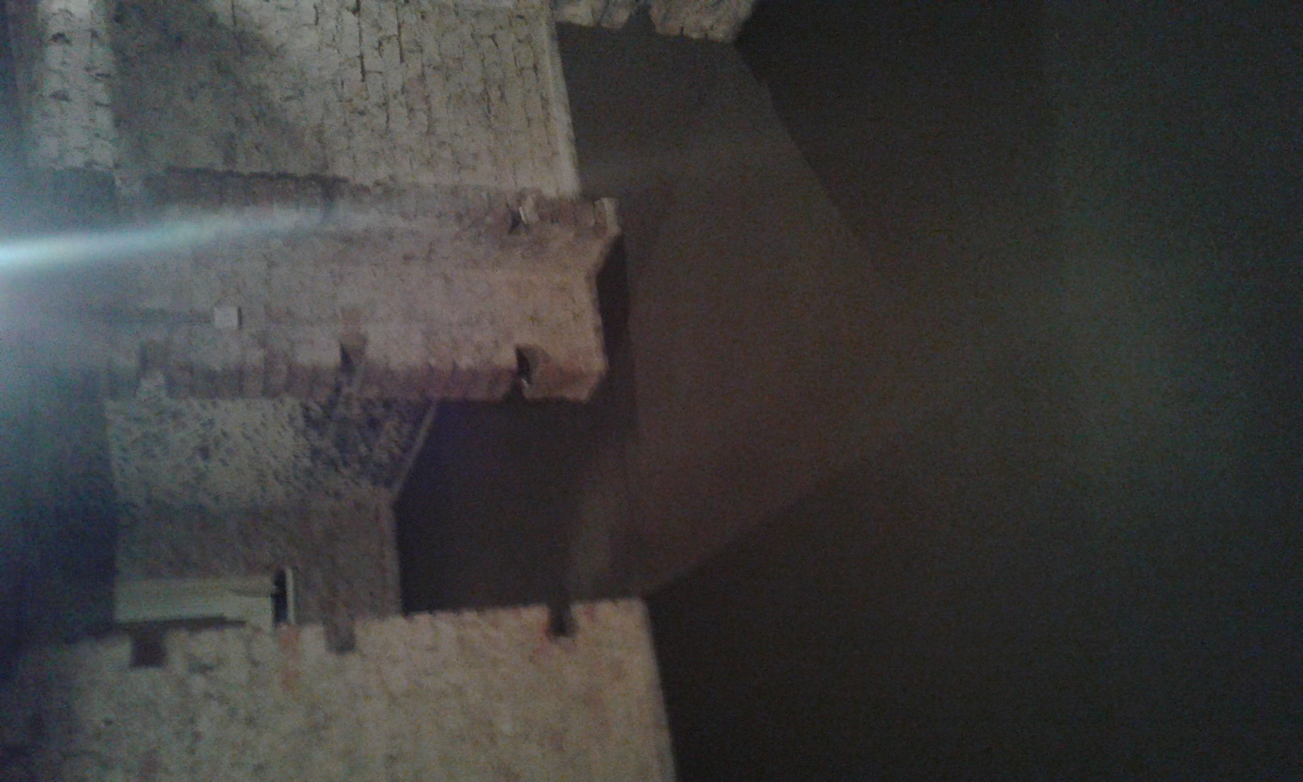 Szara, kamienna ściana z fragmentami muru, oświetlona z lewej strony, z ciemnym tłem po prawej.