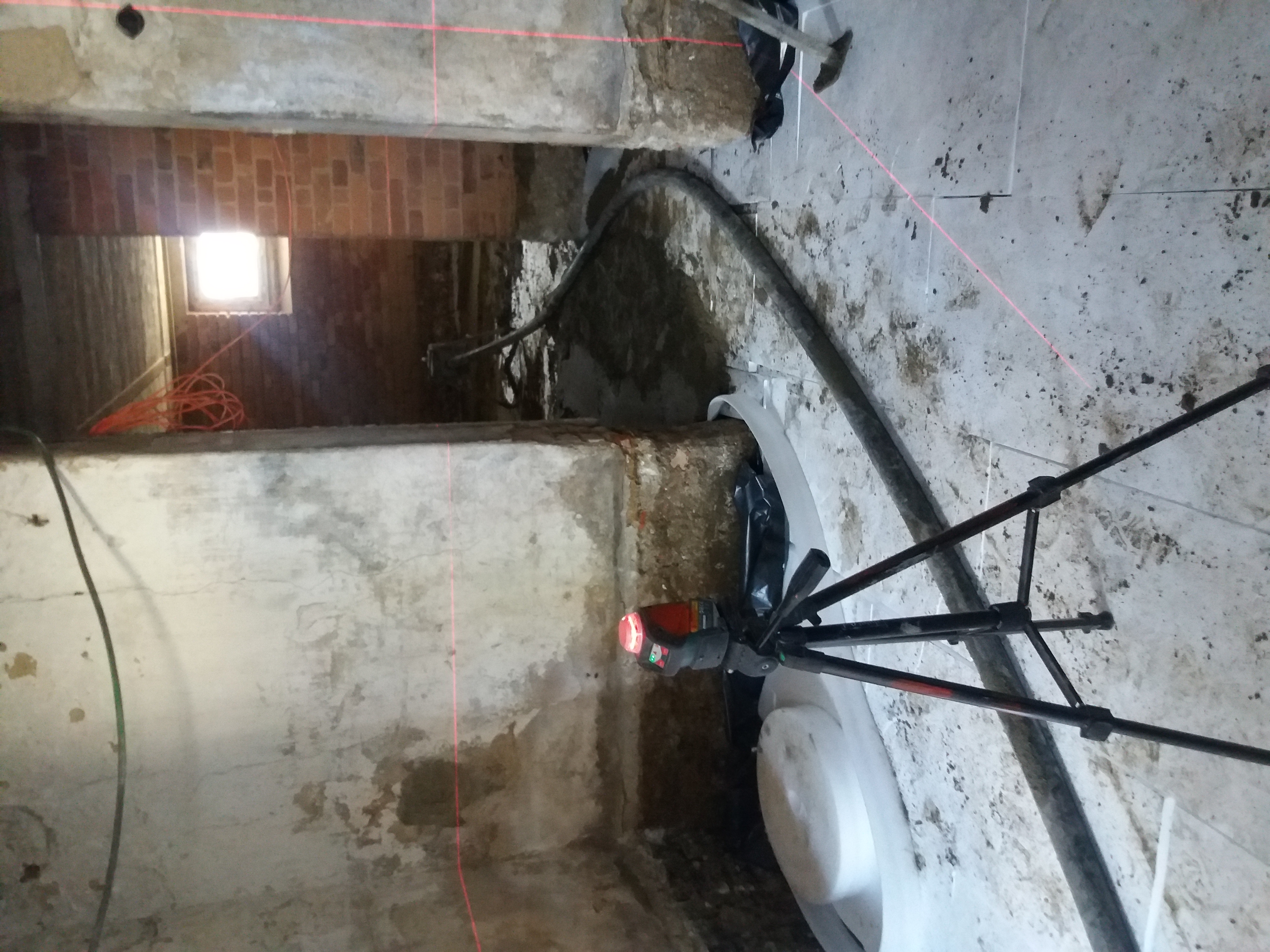 Przygotowanie podłoża pod wylewkę jastrychową: widoczny laser krzyżowy na statywie, rury doprowadzające materiał, folia izolacyjna przy ścianie oraz wyznaczone poziomy na ścianach ceglanych.