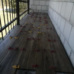 Układanie płytek imitujących drewno na balkonie, z użyciem czerwonych i żółtych klipsów poziomujących, widoczne niedokończone fragmenty posadzki.