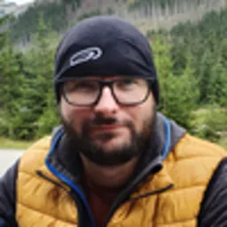 Portret mężczyzny w okularach, czapce i żółtej kamizelce na tle górskiego krajobrazu.