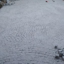 Wilewski - Szara kostka granitowa ułożona na dużej powierzchni placu przed domem, widoczny fragment budynku, przygotowany materiał do dalszych prac oraz narzędzia.