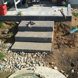 Wilewski - Trzy betonowe stopnie schodów prowadzące do tarasu z kostki, w tle widoczny dom, a obok rozrzucona ziemia i kamienie, na stopniu leżą rękawice robocze.