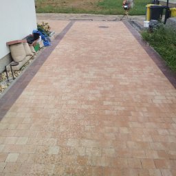 Wilewski - Nowo ułożona, beżowo-brązowa kostka brukowa na podjeździe, z widocznymi workami z materiałem budowlanym przy ścianie budynku i betoniarką w tle.