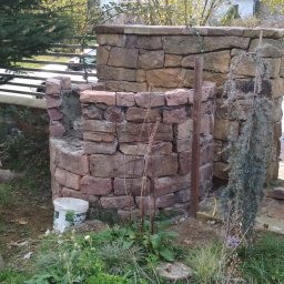 Wilewski - Konstrukcja z kamienia w trakcie budowy, widoczne elementy muru, poziomica, wiadro z zaprawą i pojemnik na szkło w ogrodzie.