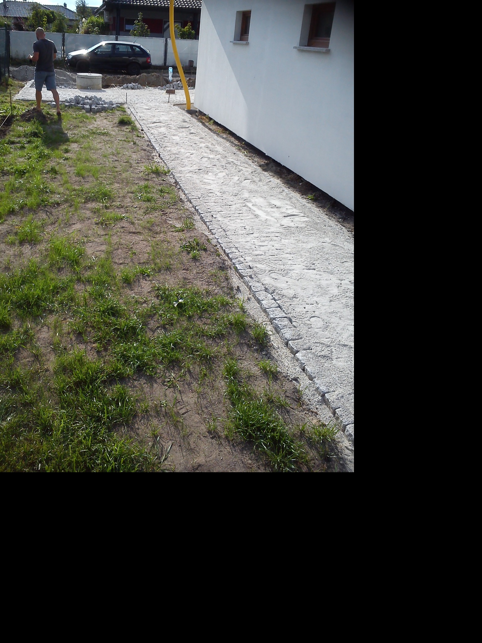 Układanie ścieżki z kostki granitowej przy białym budynku, widoczna część ułożonej nawierzchni oraz stos kostki brukowej i pracownik w tle.