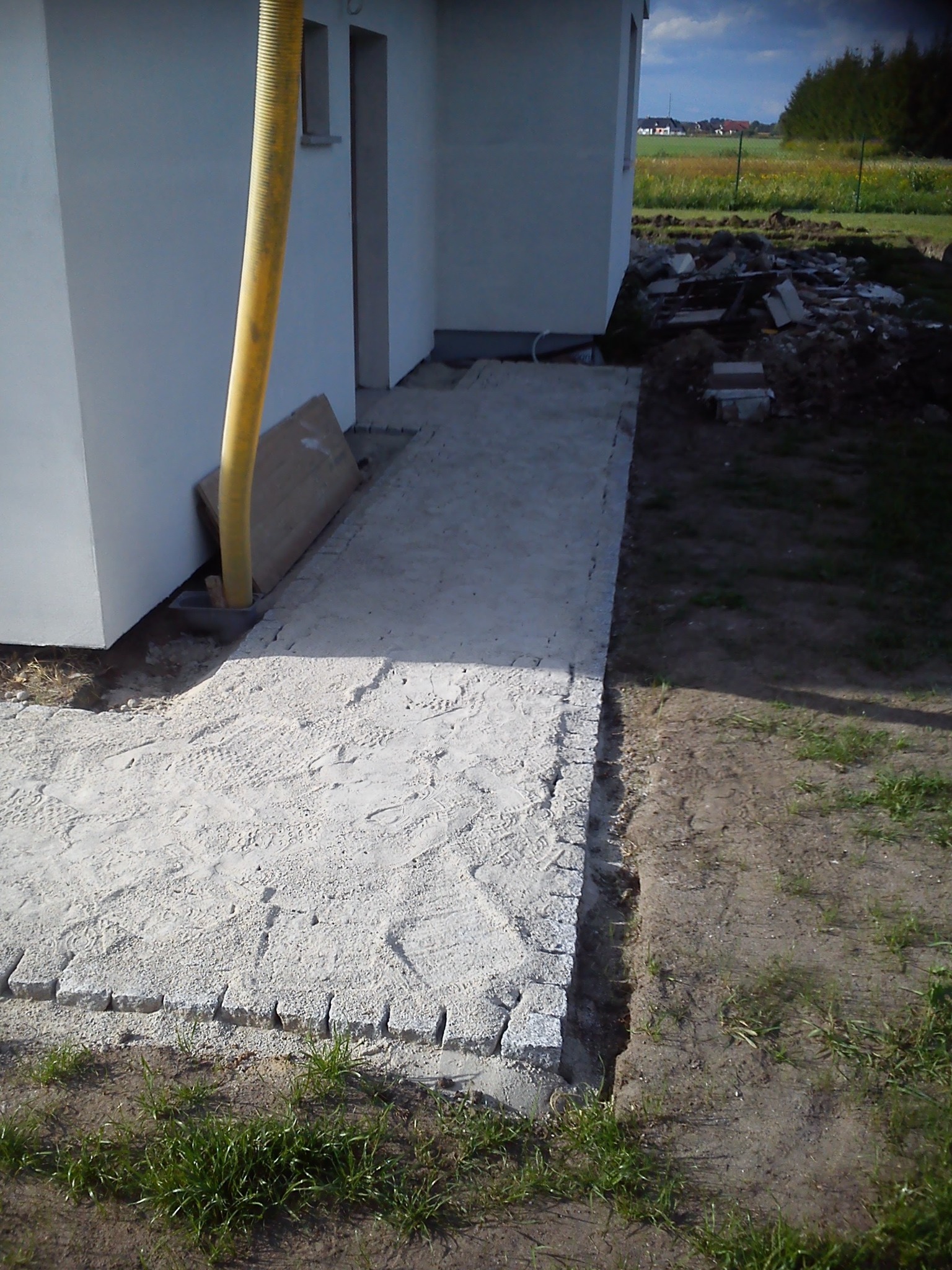 Przygotowanie podłoża pod chodnik z kostki granitowej obok białego budynku, widoczny żółty wąż wentylacyjny i ślady stóp na piasku.