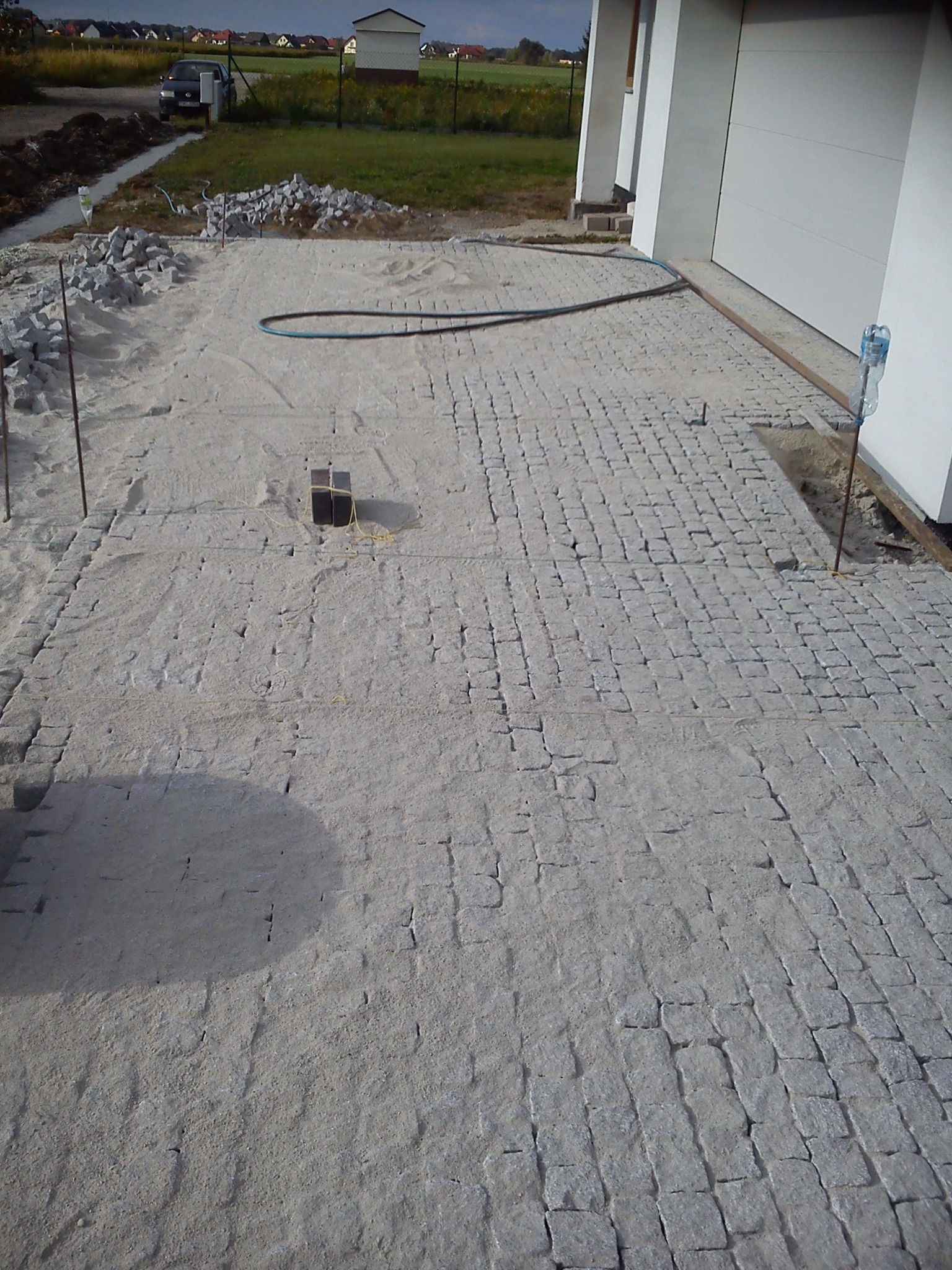 Etap prac przy układaniu szarej kostki granitowej na podsypce piaskowej przed garażem, widoczne narzędzia i materiały budowlane.
