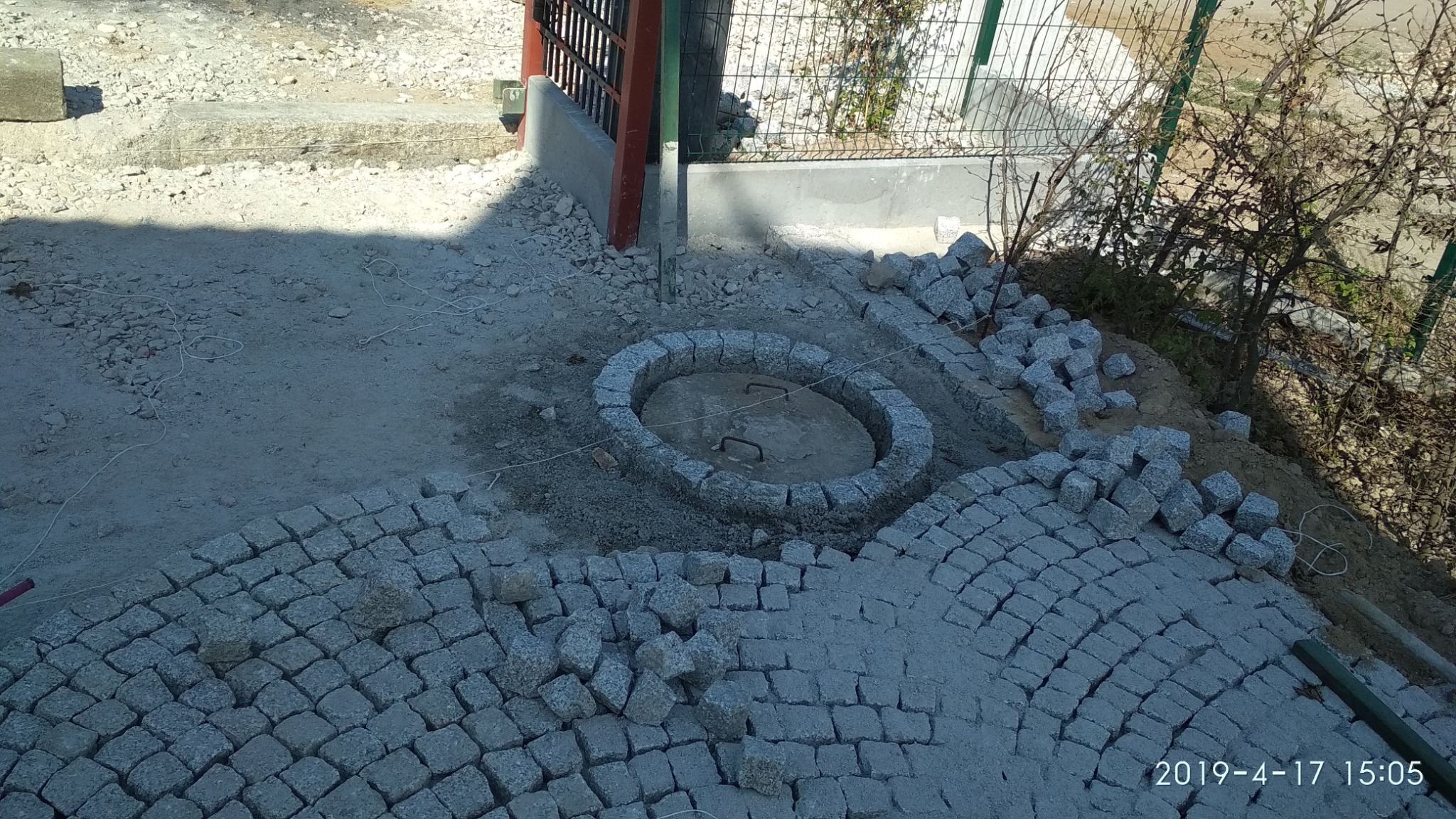 Fragment układanej nawierzchni z kostki granitowej, widoczny okrągły właz kanalizacyjny obłożony kostką, w tle ogrodzenie i ziemia.