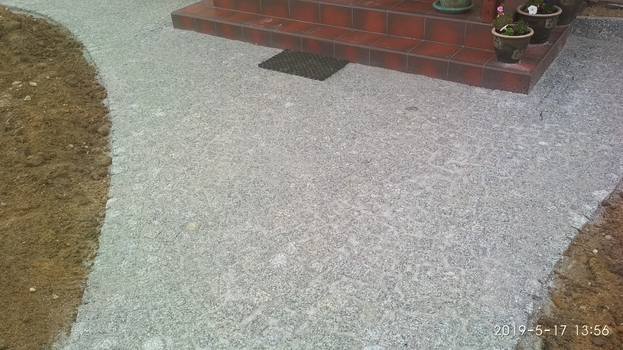 Świeżo ułożona, szara nawierzchnia z drobnego kruszywa przy schodach wejściowych do domu, z widocznym pasem ziemi po lewej stronie.