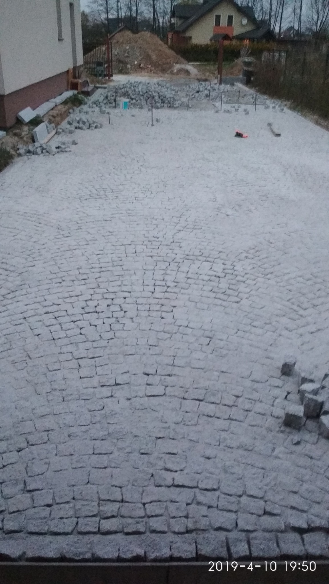 Szara kostka granitowa ułożona na dużej powierzchni placu przed domem, widoczny fragment budynku, przygotowany materiał do dalszych prac oraz narzędzia.