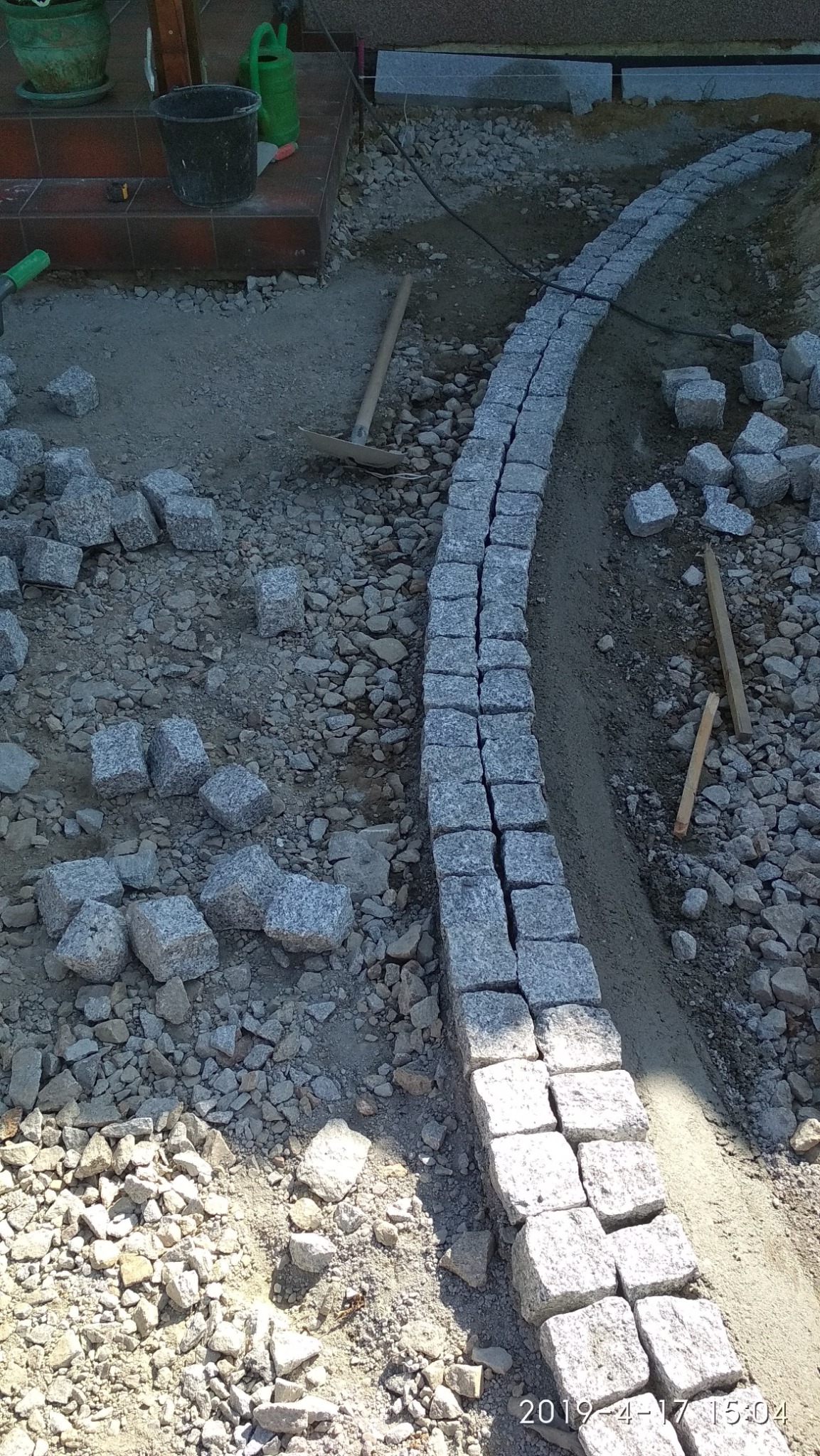 Wykop pod obrzeże z kostki granitowej, widoczne przygotowane podłoże, kostka ułożona w łuk, narzędzia pracy i luźne kostki obok.
