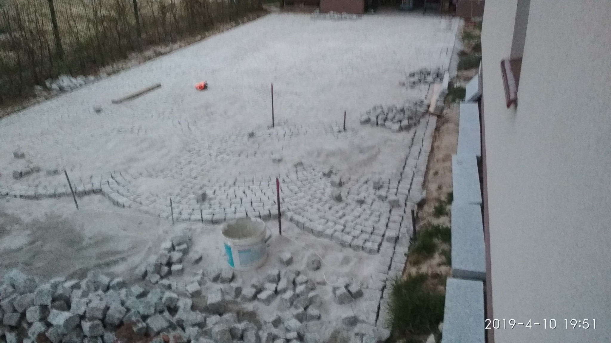 Układanie kostki granitowej na posesji, widoczny fragment ułożonego wzoru w łuk, przygotowane kamienie, wytyczone granice i wiadro z zaprawą.