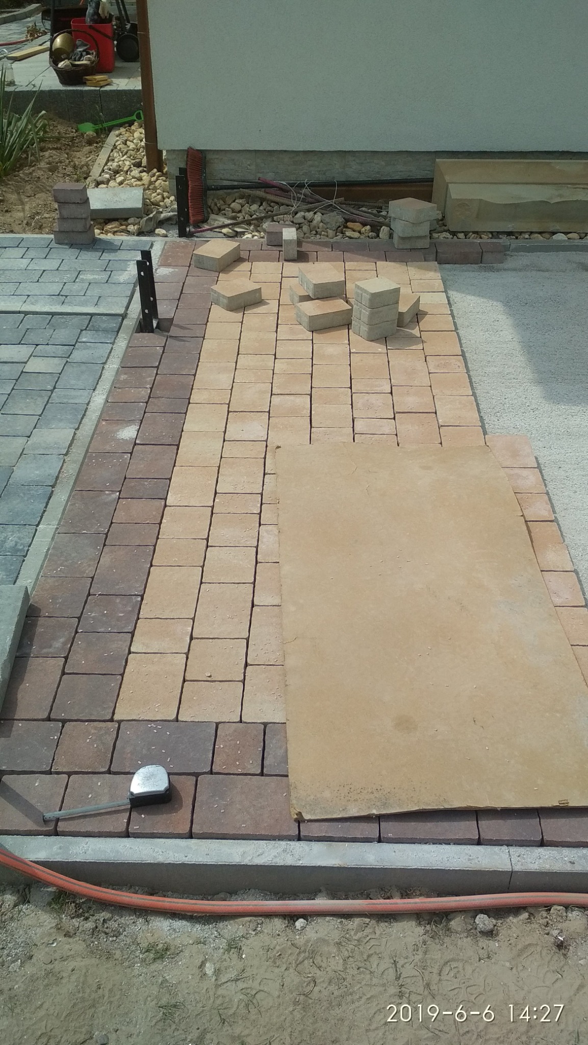 Fragment układanej nawierzchni z kostki brukowej w odcieniach brązu i beżu, widoczne narzędzia pomiarowe, płyta ochronna i przygotowane stosy kostki.