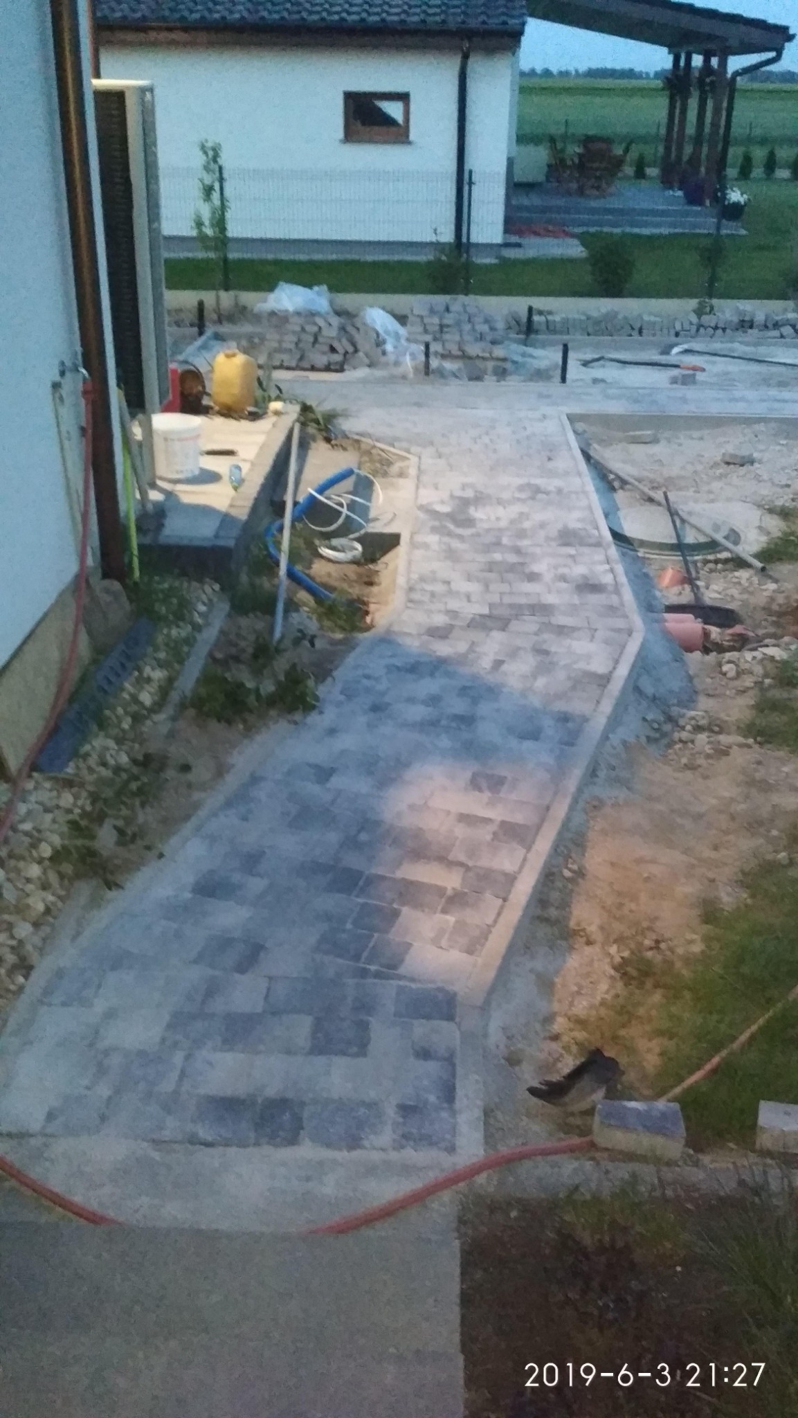 Wykładanie ścieżki z szarej kostki brukowej w ogrodzie, widoczne narzędzia budowlane i zapas kostki