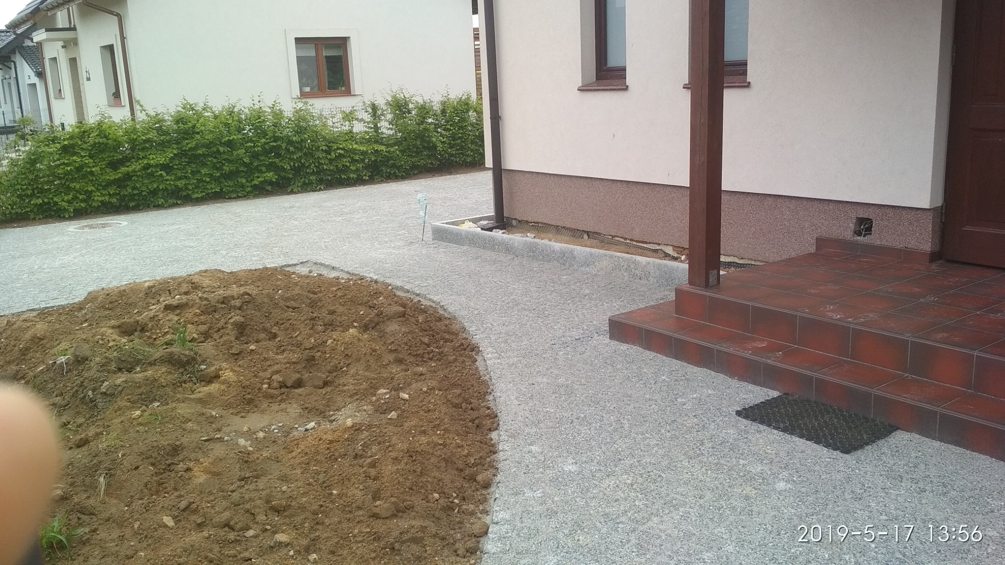 Świeżo ułożona, szara nawierzchnia z drobnych kamieni prowadząca do wejścia domu z terakotowymi schodami, obok widoczna pryzma ziemi.