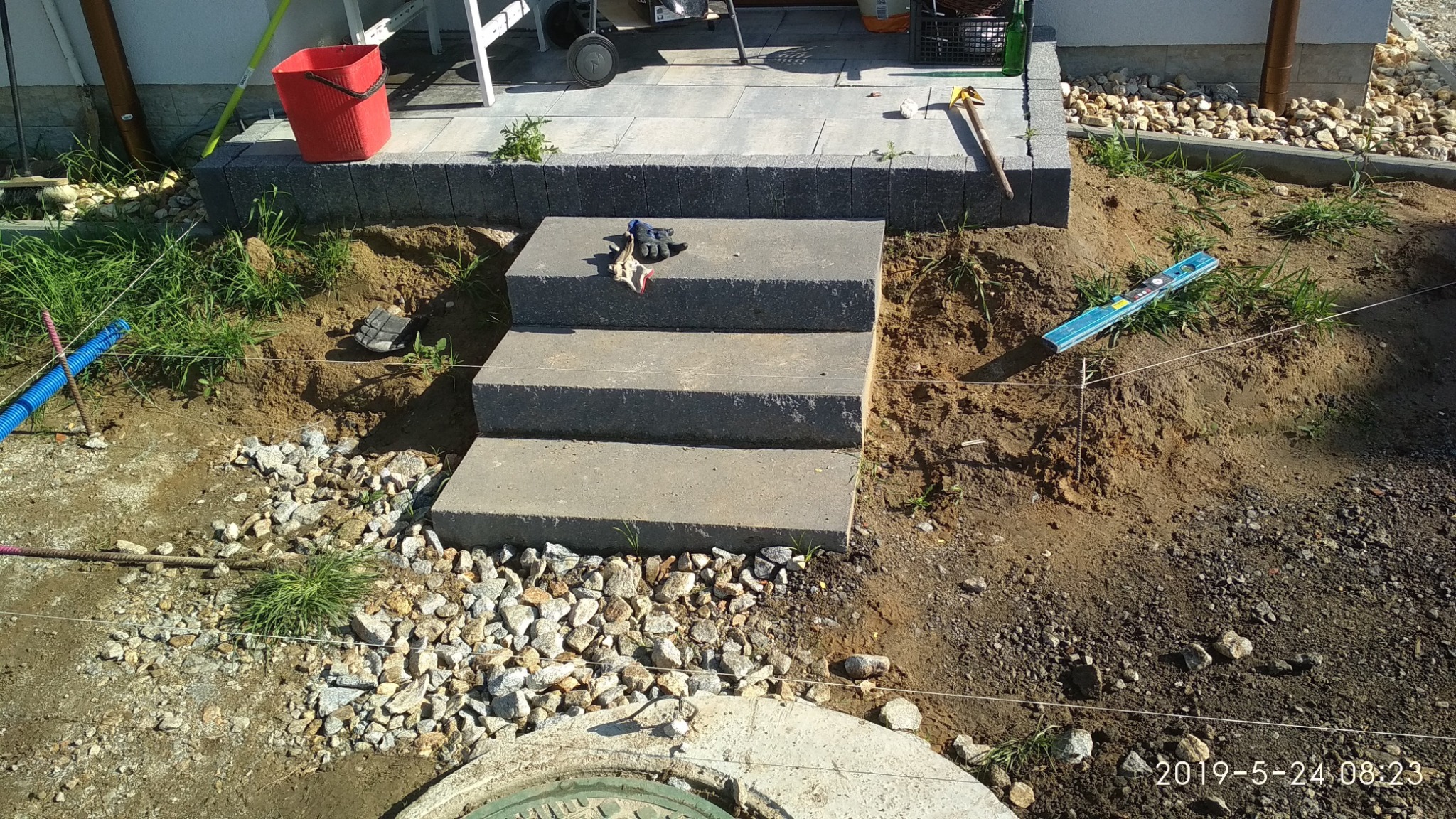 Trzy betonowe stopnie schodów prowadzące do tarasu z kostki, w tle widoczny dom, a obok rozrzucona ziemia i kamienie, na stopniu leżą rękawice robocze.